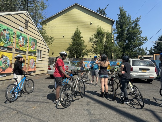 Seven high school-aged youth gather in a parking lot facing an adult instructor, all with bikes.