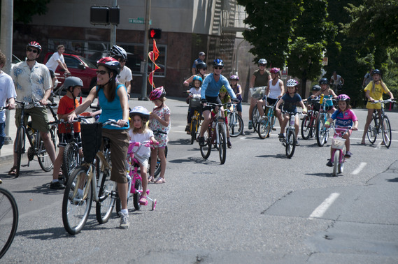 Dozens of adults and children ride bikes on a downtown Portland street on a sunny day.