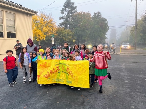 More than 20 elementary students huddle around a large yellow paper banner, "International Walk & Roll to School Day James John"