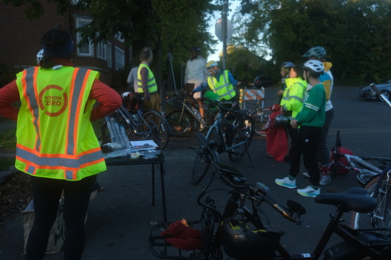 About nine adults wearing high-visibility gear huddle around before a bike ride as the sun starts to set. 
