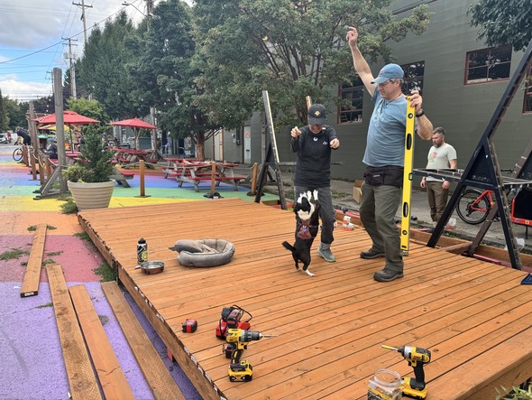 Two men stand atop a wooden stage being built at Ankeny Rainbow Road. They take a moment to play with a dog.