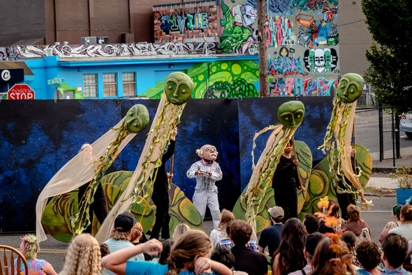 A street performance features large green puppet heads and a person in an astronaut costume in front of a painted backdrop