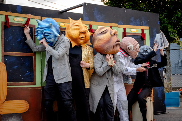 Performers wearing oversized, colorful puppet heads act out a scene in front of a backdrop resembling a train.