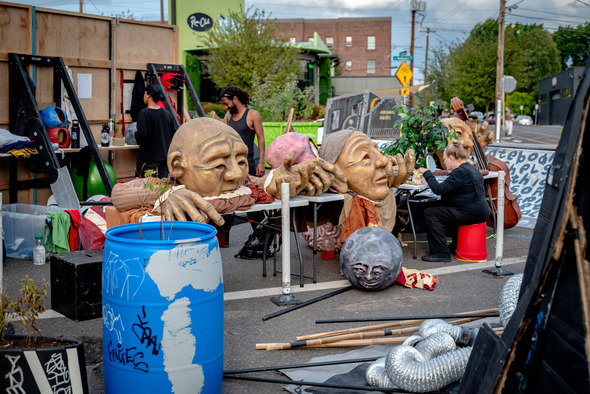 Performers prepare for a show, setting up large puppet heads and props on tables behind stage.