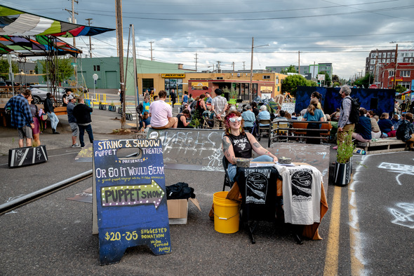 A person sits at a booth for the puppet show,. attendees gather in the background for the outdoor performance.