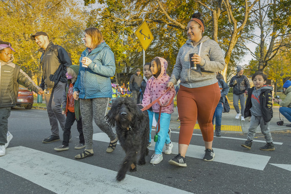 A group of adults and elementary school students walk across a crosswalk in front if a school in the fall.