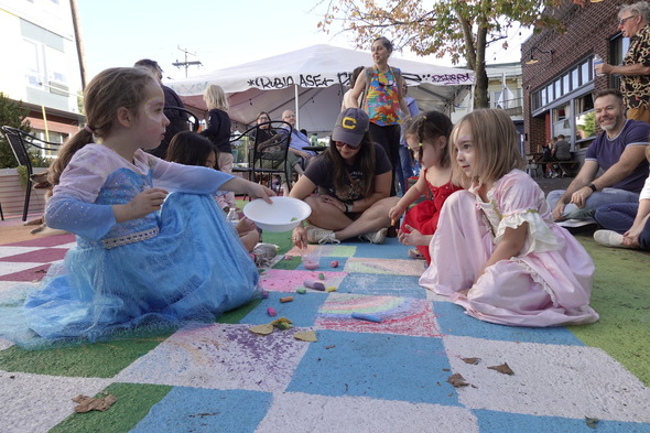 Children sit in the plaza and draw with chalk on the colorfully painted street. Adults supervise and mingle in the background.