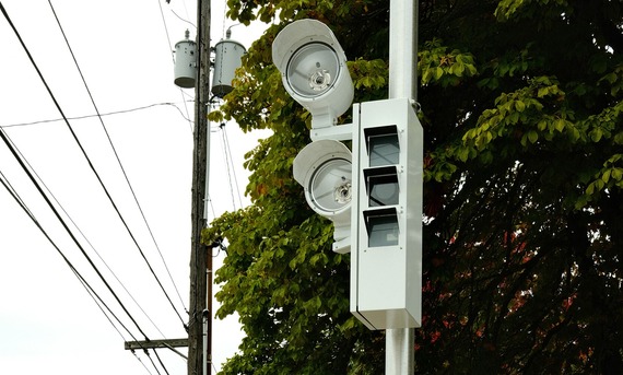 A vertical, white traffic safety camera has multiple flash bulbs and lenses and is in front of tree foliage. 