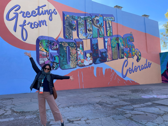 Safe Routes to School staff, Jeri Stroupe, stretches her arm wide in front of a "Greetings from Fort Collins, Colorado" mural.