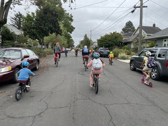 About a dozen adults and children bike and scoot down a neighborhood street on their way to school.