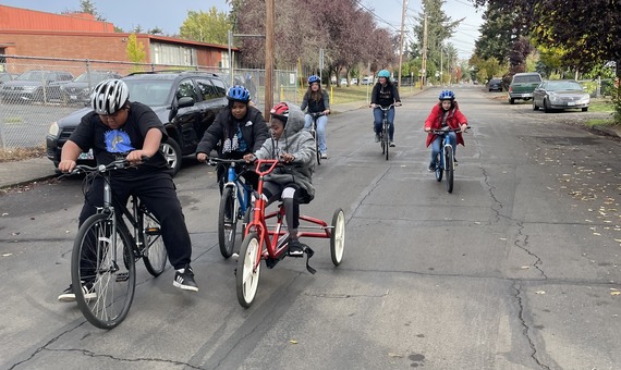 Six students rides different styles bikes, including one adaptive bike, along a neighborhood street next to their school. 