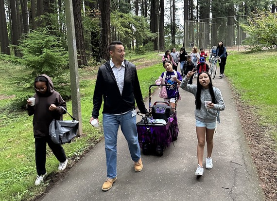 A group of students and adults walk to school together through a park on a paved path.