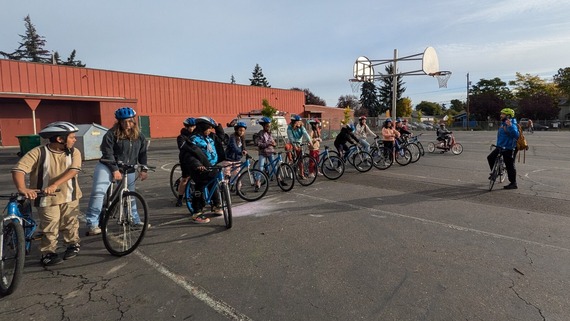 Elementary school students stand with their bikes in a line on a schoolyard listening to their teacher's instructions.