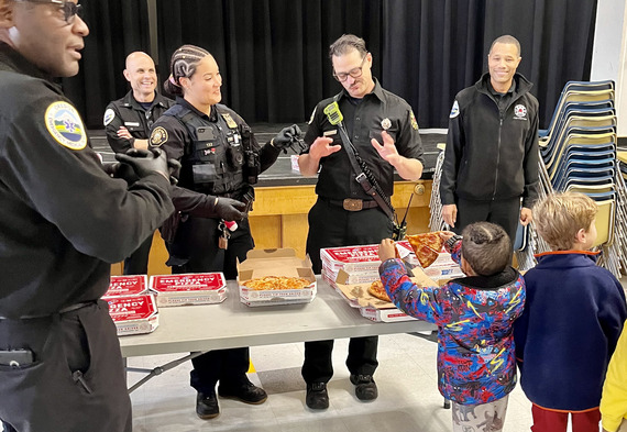 Several Police and Fire and Rescue personnel in uniform hand out pizza to elementary school students.