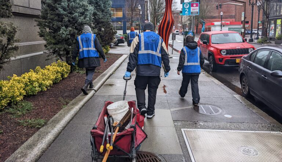 Several people in safety vests walk along a city street, pulling a wagon with supplies for picking up trash