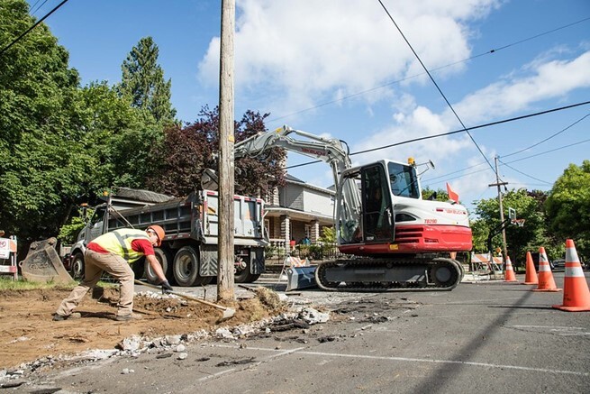 Excavator disposing dirt inside a dump truck and construction worker raking dirt where concrete was removed