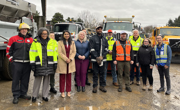 Priya Dhanapal and people in safety vests, stand in a muddy lot in front of heavy equipment for a PBOT and agency partners press conference