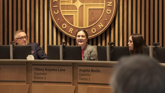 A photograph Councilor Koyama Lane in chambers. She's in between Councilors Ryan and Morrillo. Behind them are vertical wood slats of multiple shades