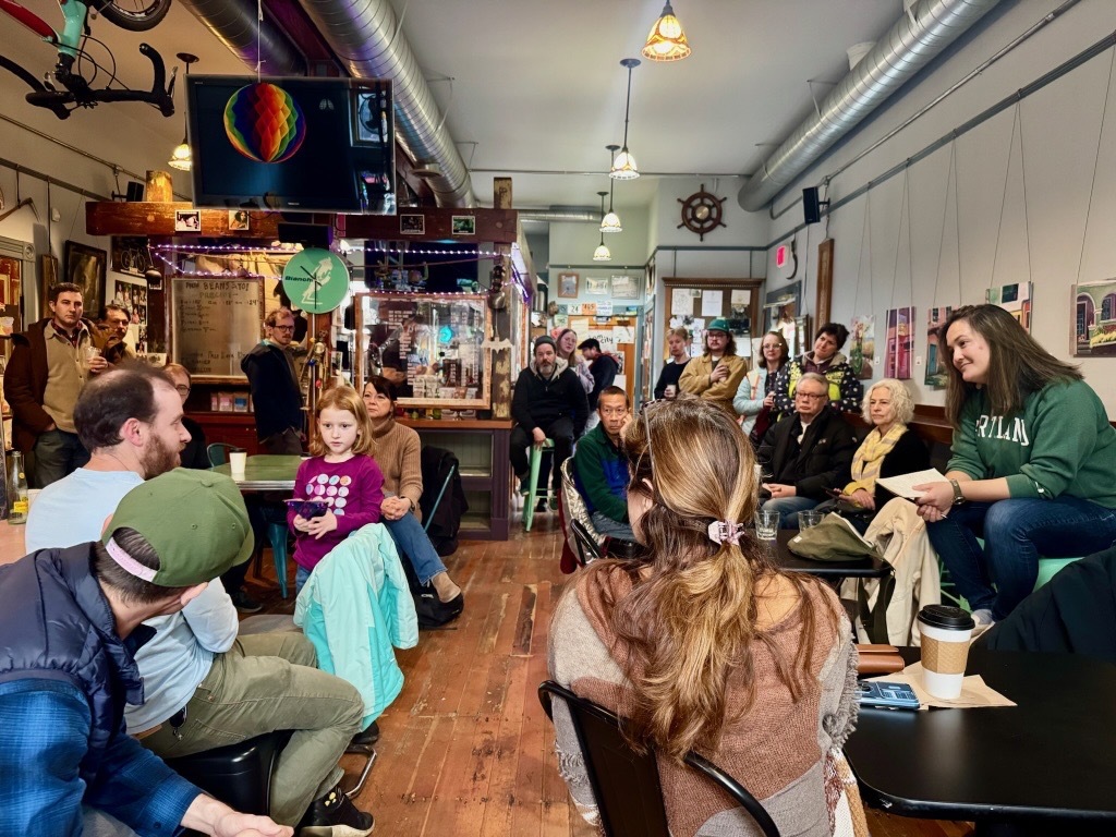 Councilor Koyama Lane sitting on the right listening, with people all around in a coffee shop