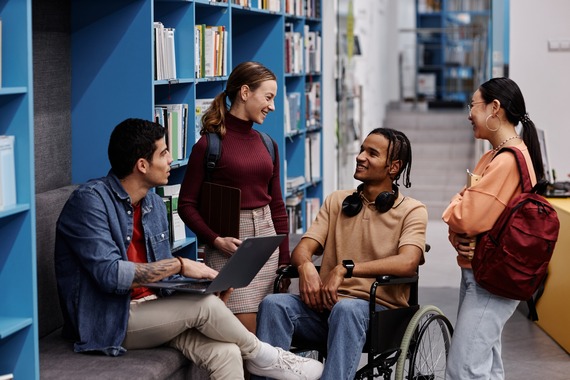Young adult students in college library talking with black man in wheelchair