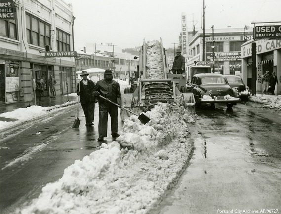Two people with shovels stand in the middle of a city street that is half covered in a piles of snow and half cleared, while next to a truck