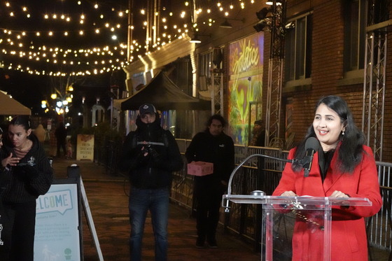 Priya Dhanapal stands at a podium in front of Ankeny Alley, pedestrians pass through, overhead lighting hangs above.
