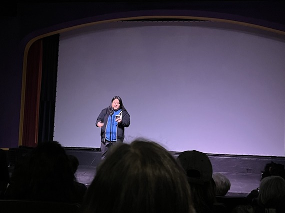Woman on stage in front of purple-lit dark screen reading to audience