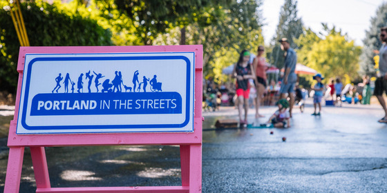 A pink street barricade with the logo for Portland in the Streets. In the background a crowd gathers for an event in the street.