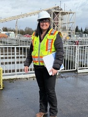 Engineer Christa Overby stands outside a wastewater treatment plant, wearing a hardhat and holding a clipboard.
