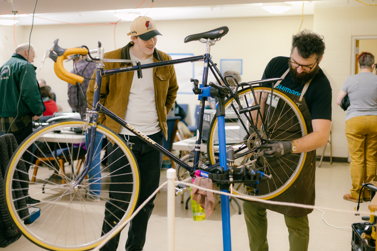 A bike mechanic works on a bike in a stand while the grateful owner observes.
