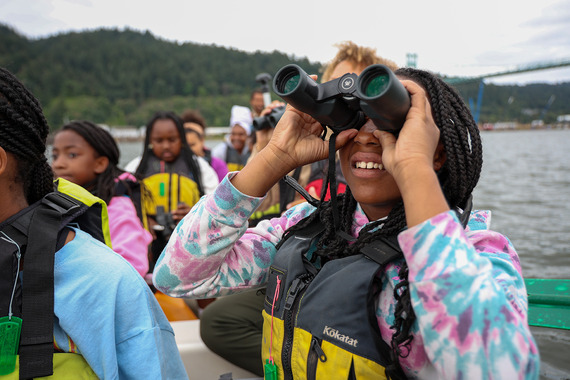 Students and adult staff wear lifejackets while sitting in a canoe. A student in the front row looks through binoculars. 