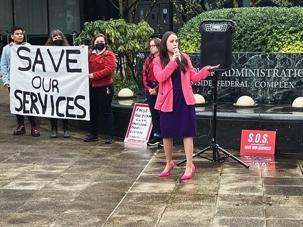 Councilor Morillo speaks to a crowd. In the background advocates are holding a sign that says "SAVE OUR SERVICES"