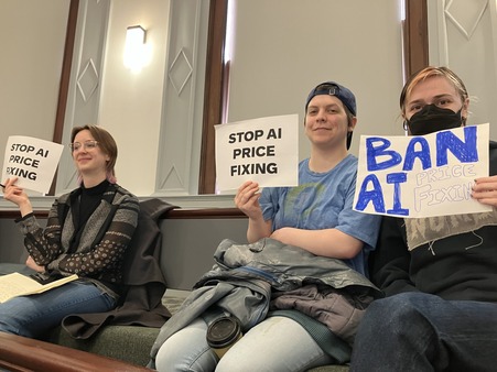 Advocates sit in Council chambers holding signs that say "stop AI price fixing"