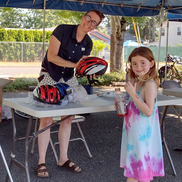 A smiling adult stands behind a table outside, holding a bike helmet, as a smiling child stands in the foreground. Transportation ambassador