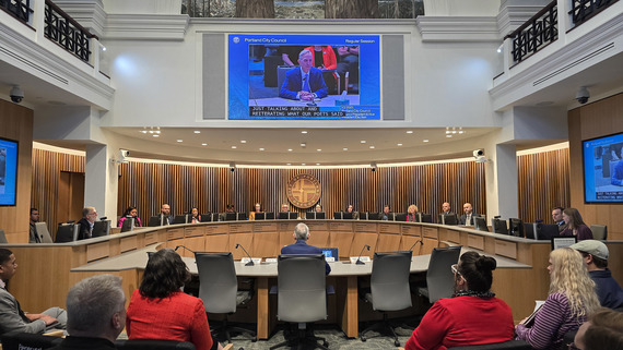 Portland City Council chambers with the 12 city councilors seated in a U-shape and a speaker seated facing them