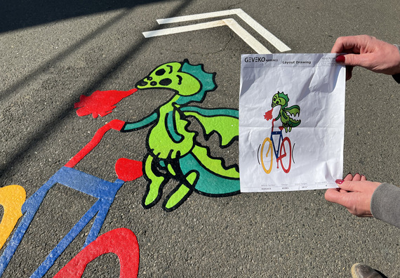 Bike lane art shows a dragon riding a bicycle with a pair of hands holding the paper artwork showing the same image