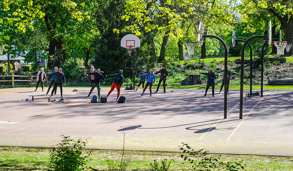 FItness in the Park at Irving Park, where about a dozen people follow an exercise instructor while working out on the basketball court at a city park.