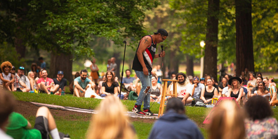 A man speaks holding a microphone in the middle of a seated crowd with smiling people in the middle of a city park for Comedy in the Park
