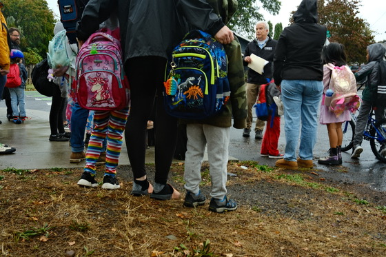 A group of kids and adults stand on a street corner on their way to school on a rainy morning.