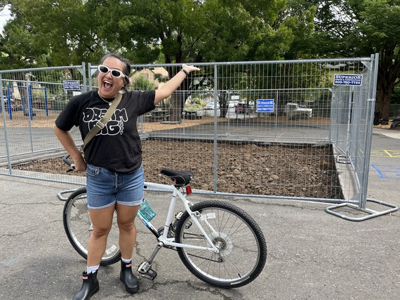 Councilor, wearing sunglasses and standing with her bike, gesturing toward a fenced-in rectangle of soil cut out of schoolyard asphalt