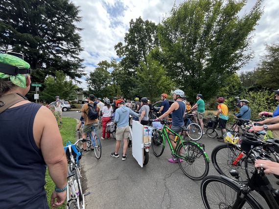 Many bicyclists standing next to their bikes, listening to a speaker with a megaphone