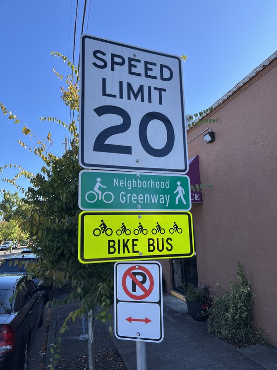A white 20 mph speed limit street sign above a green “neighborhood greenway” sign above a yellow “bike bus” sign above a no parking sign.