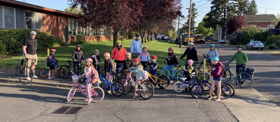 18 elementary school kids and adults arrive to school with their bikes on a sunny day.