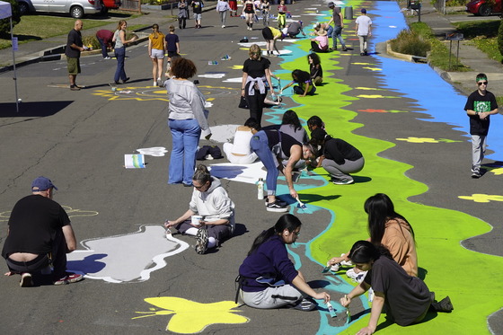 More than 30 teenagers and adults paint a street art mural using green, yellow, blue, red, and white colors on a street.