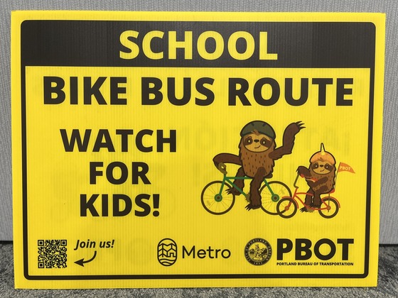 A yellow sign with black text, "School Bike Bus Route Watch For Kids!," as well as an illustration of two sloth characters on bikes.