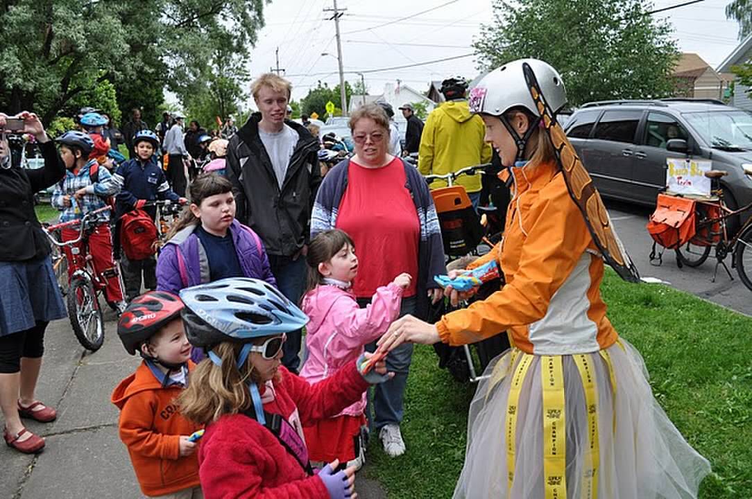 An adults wearing butterfly wings and a tutu hands incentives to young children on a sidewalk.