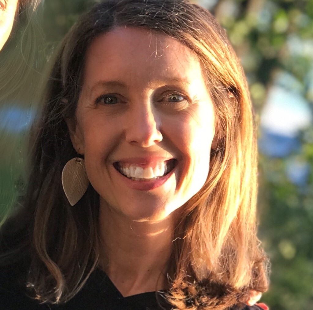 A headshot of a woman wearing leaf earrings and smiling as sun reflects off the left side of her face. 