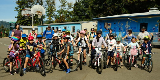 About 30 elementary school kids and adults stand with their bikes on a school basketball court on a sunny day.