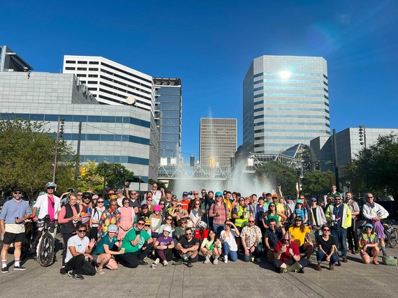 Cyclists pose for camera in front of Salmon Springs Fountain in downtown Portland on sunny day.