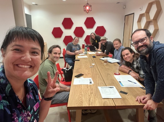 Councilor TKL smiling at the camera and showing the peace sign in front of seven people gathered around a table and smiling at the camera